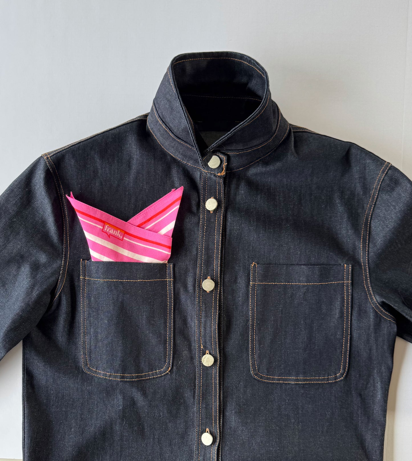 Dark denim jacket with a pink & white striped handkerchief inside the pocket on a light gray background