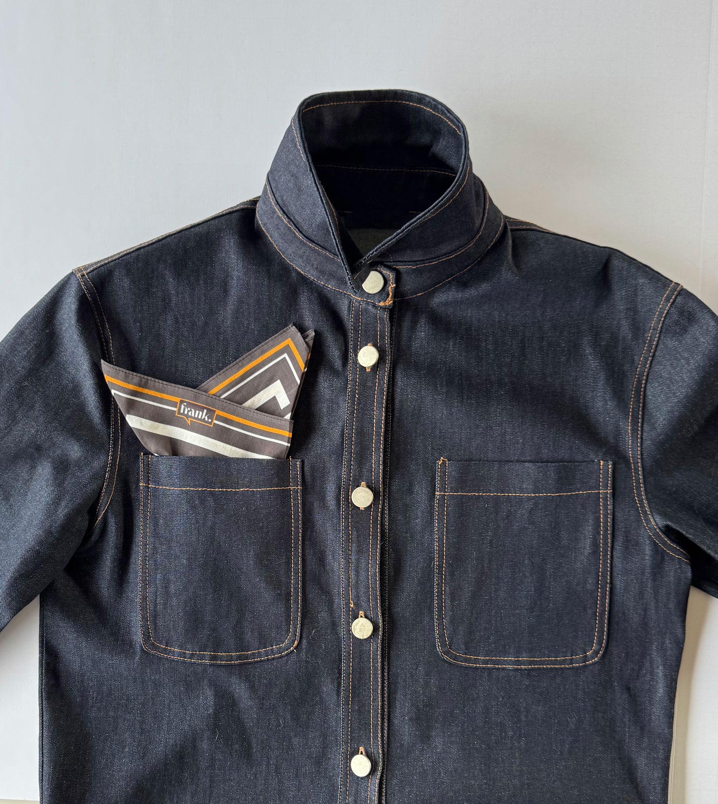 Dark denim jacket with a black and gold striped handkerchief tucked into the pocket on a light gray background