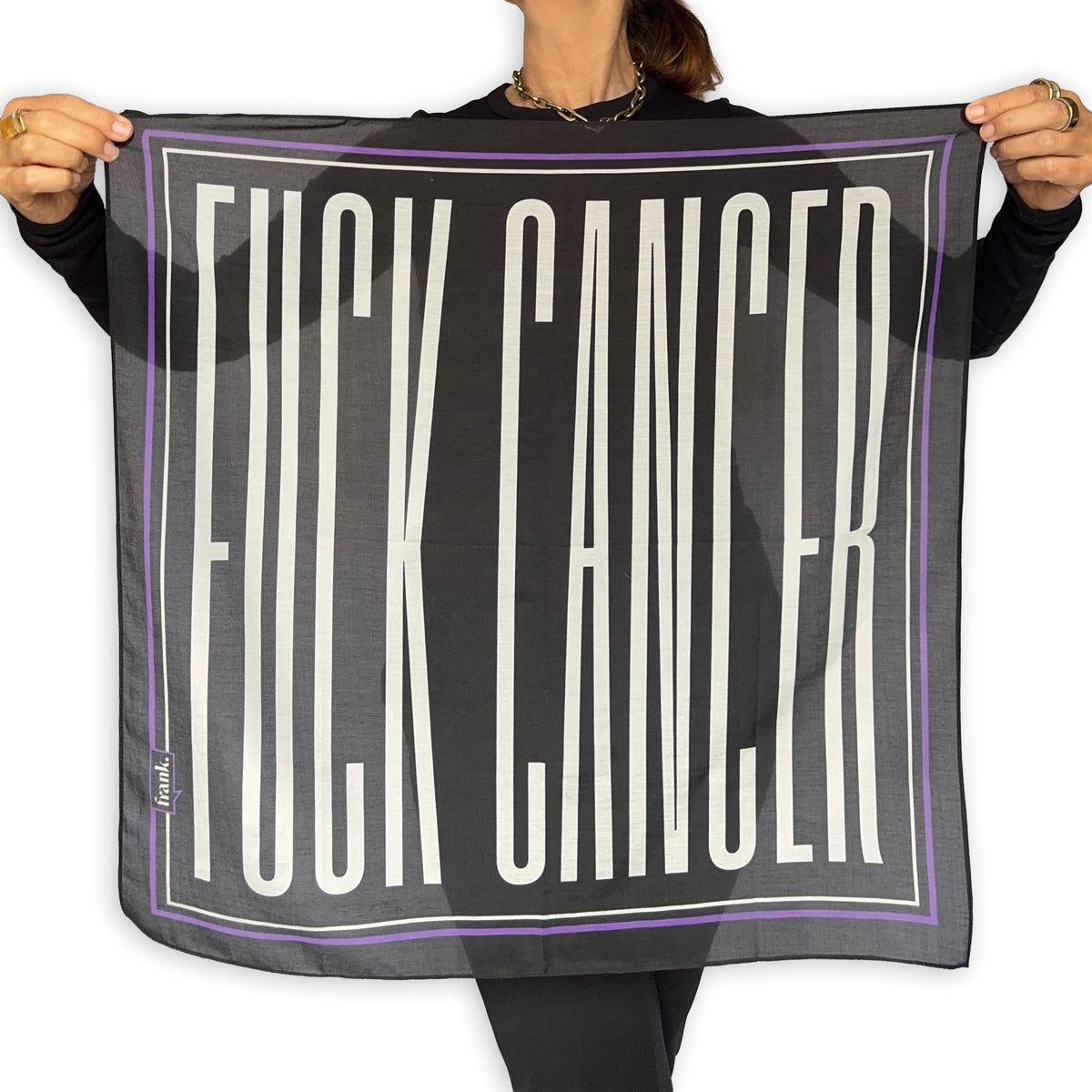A person holds up a square scarf with the words 'FUCK CANCER' printed in large white letters on a black sheer background, bordered by thin white and purple lines. The person is wearing a black long-sleeve top and black leggings against a plain white background.