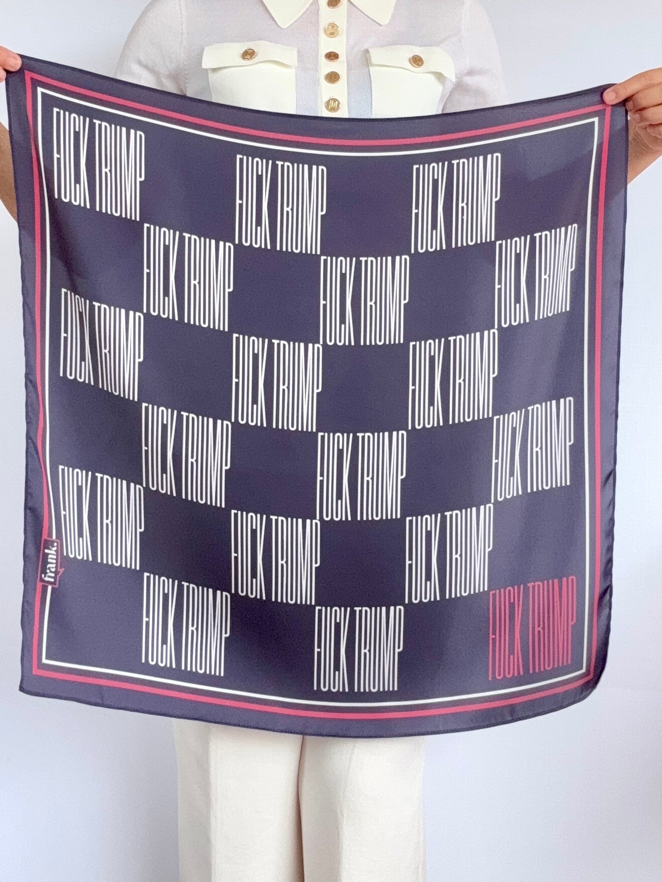 Woman holding navy scarf with a checkerboard pattern of 'F*ck Trump' text on a blue background with a red border. 