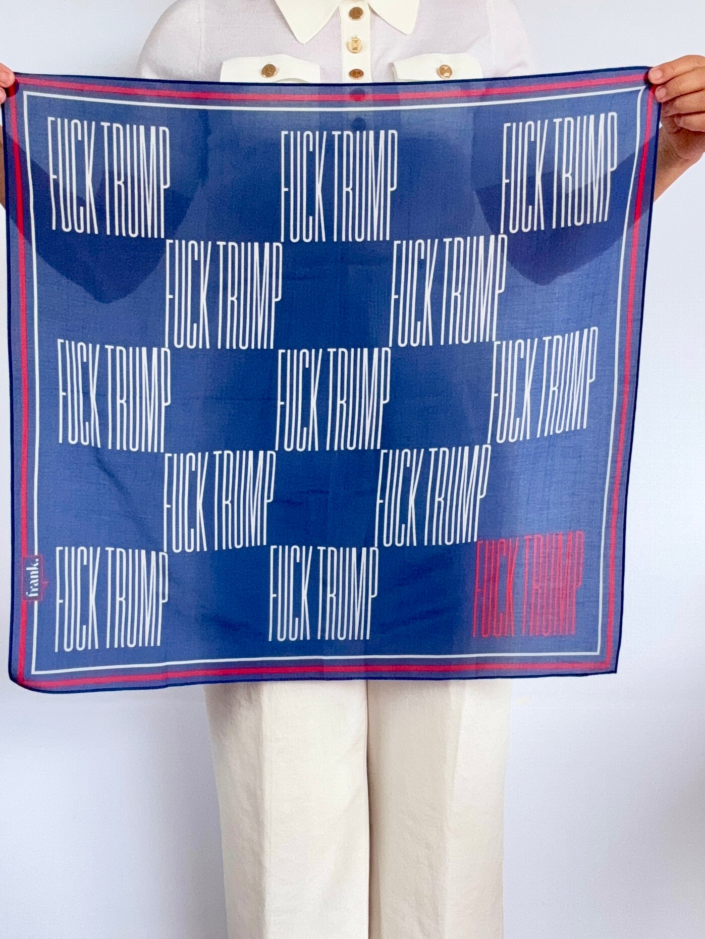 A person holds up a square scarf featuring the text 'FUCK TRUMP' repeated in white on a dark blue background, with a red and blue border and one red word in the lower right. The person is wearing a white collared shirt and white pants against a light gray background.