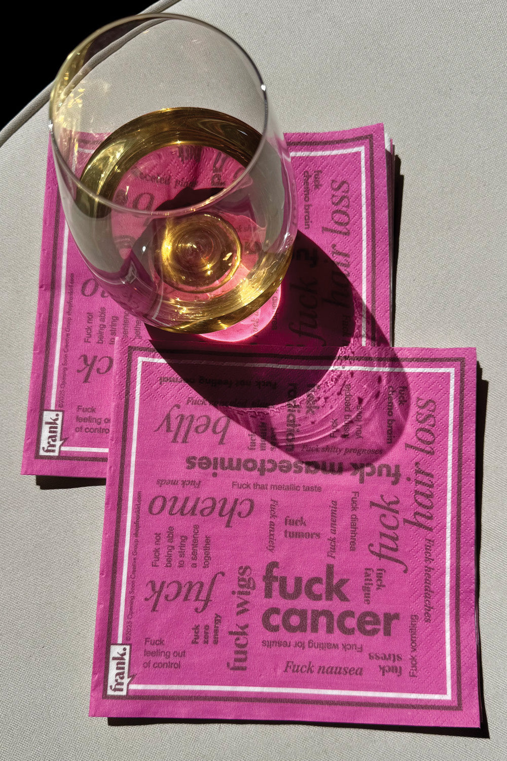 Hot pink and burgundy Fuck Cancer Cocktail napkins sitting on an outdoor table with a glass of white wine.