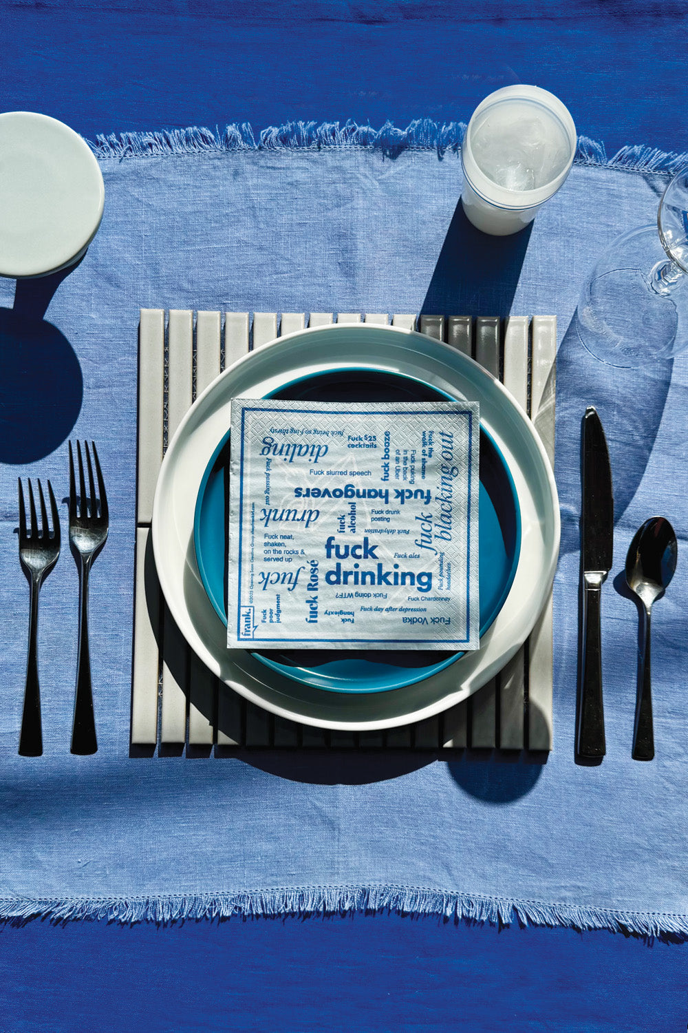 Table setting with a blue placemat, white plates, black cutlery, and a Fuck Drinking cocktail napkin on a plate.