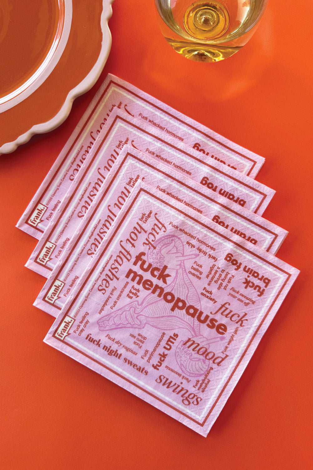 Fuck Menopause pink and orange cocktail napkins fanned out on an orange table top