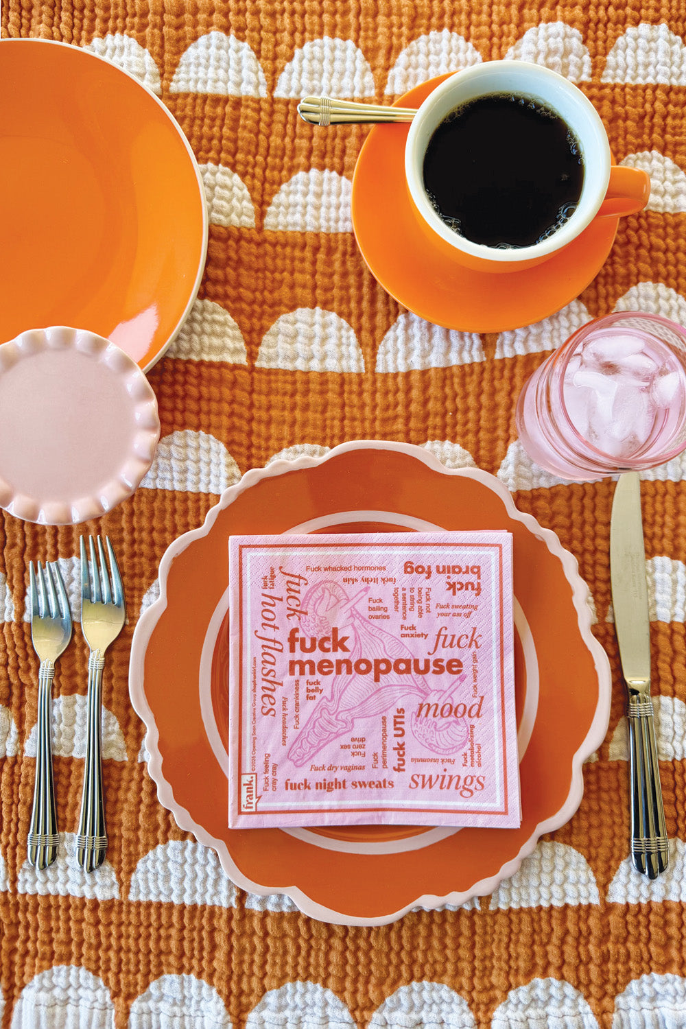 Table setting with a pink Fuck Menopause cocktail napkin sitting on orange plates, and a cup of coffee on an orange tablecloth.
