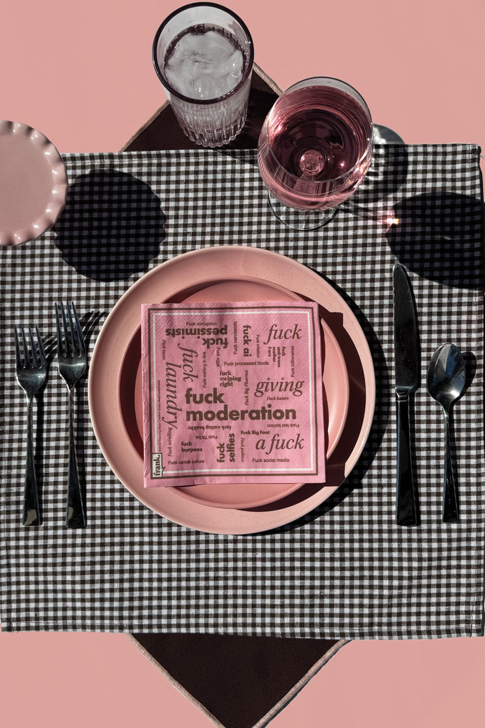 Table setting with pink plates, black cutlery, and a checkered tablecloth on a pink background. The Fuck Moderation cocktail napkin is sitting on dinner plate.