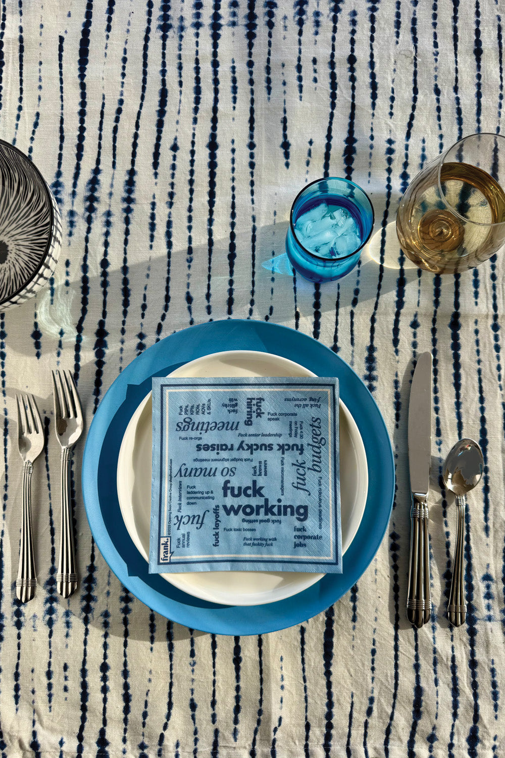Table setting with a blue plate, white plate, Fuck Working blue cocktail napkin, and glass of white white on a striped batik tablecloth.
